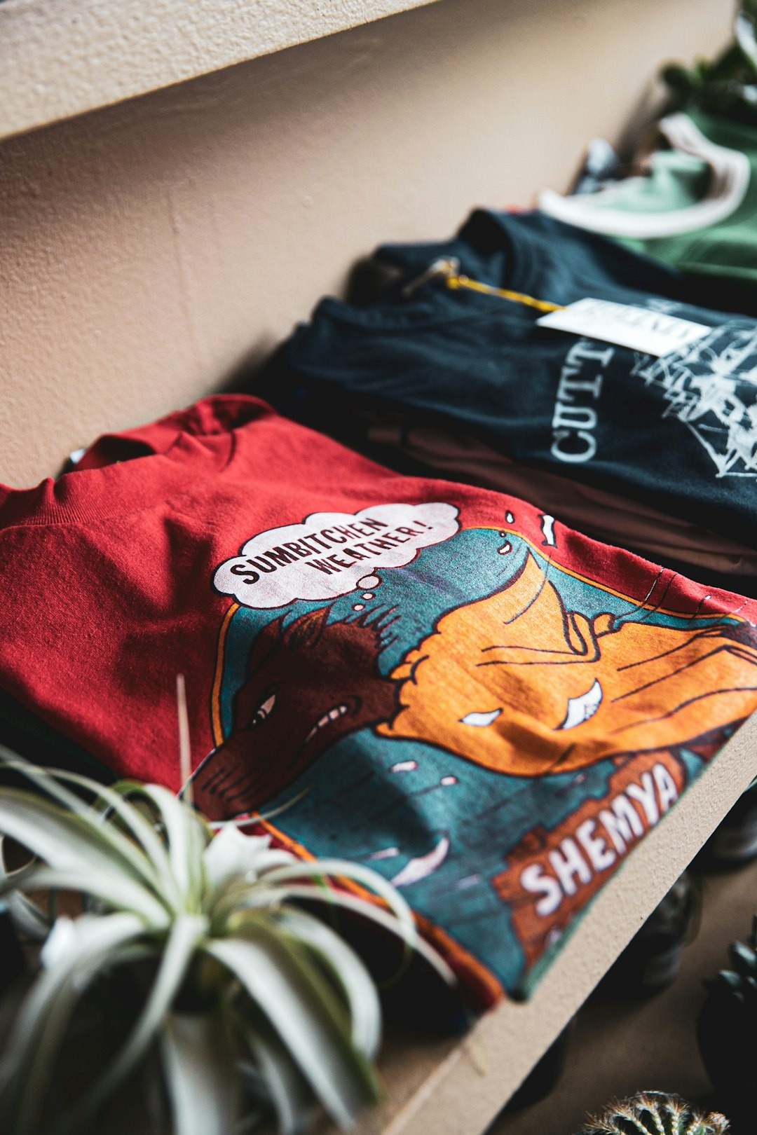 Red t-shirt with 'SUMBITCHEN WEATHER!' graphic on a wooden shelf.